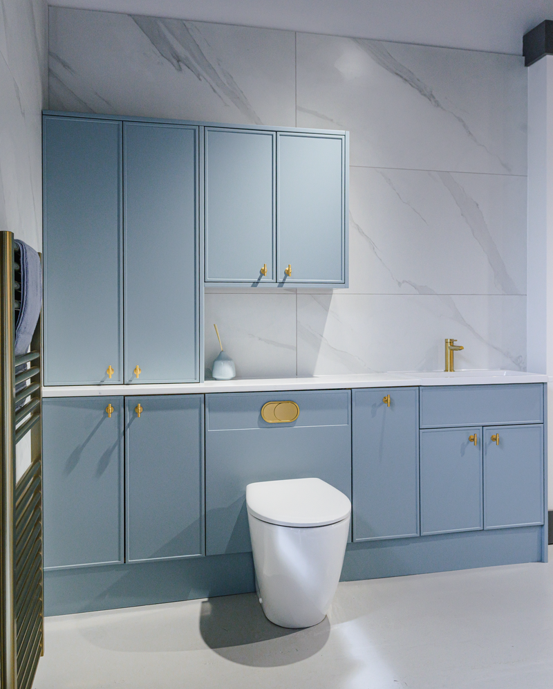 Oceanair Perth Showroom Bathroom Design, baby blue wall bathroom cabinets, concealed white toilet, gold handles and taps, white sink, white and grey marble-effect large format wall tiles, bronze heated towel rail with grey hand towel, white resin floor.