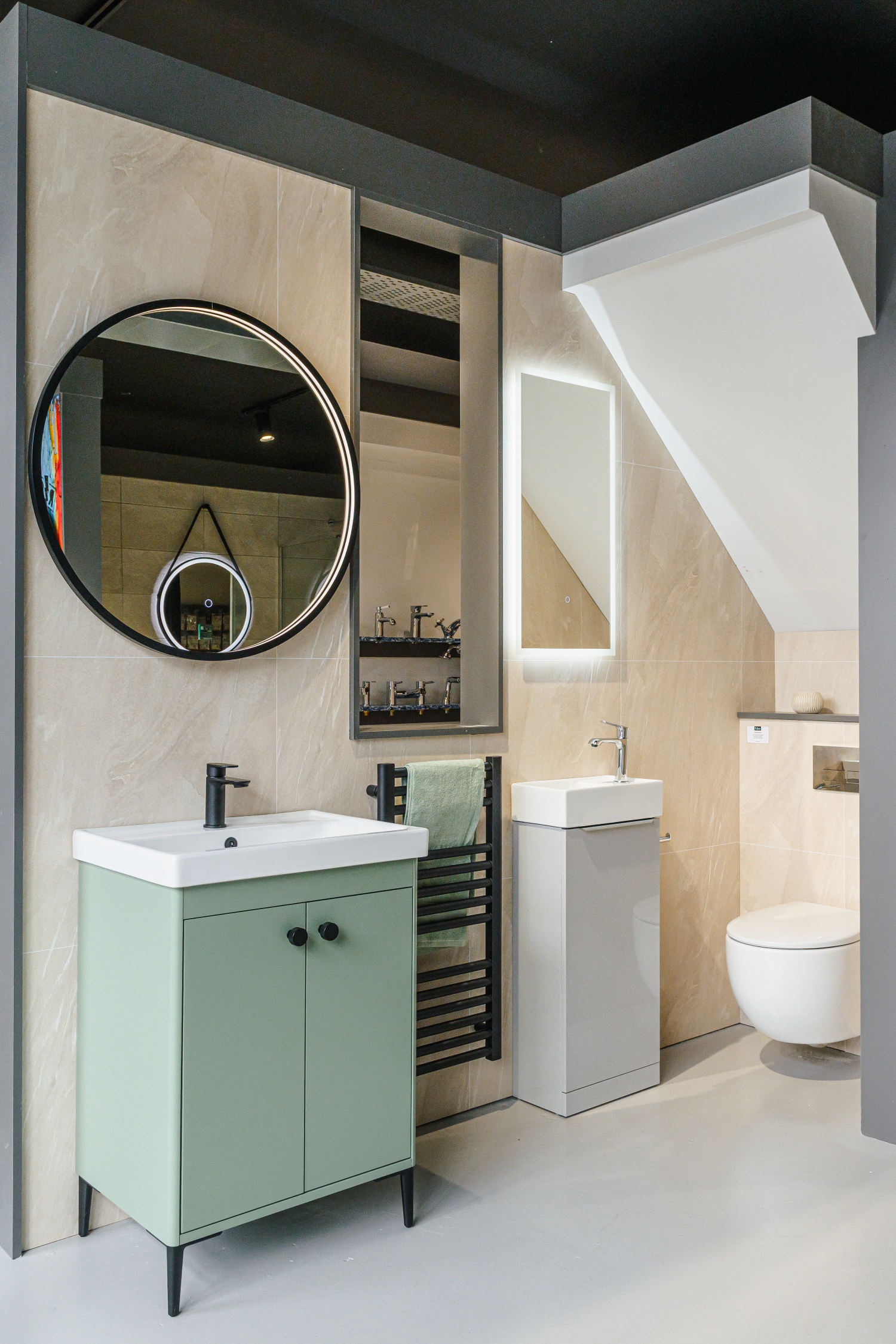 Oceanair Perth Bathroom Showroom Display. sage green freestanding under basin cabinet, rectangular basin with black mixer tap. Small black heated towel rail, concealed cistern toilet under sloping ceiling. Lit circular mirror over basin. Beige marble-effect large format wall tiles. Vertical rectangular mirror. Backlit vanity mirror.